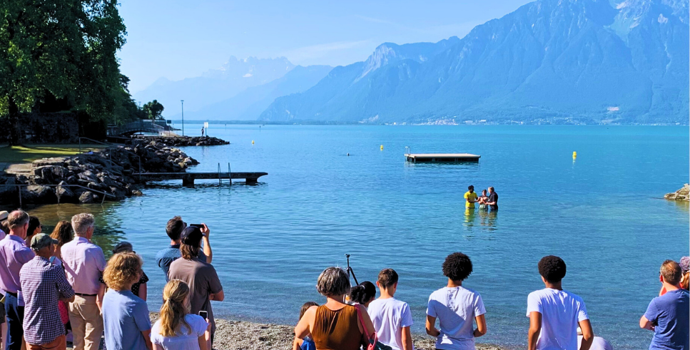 Baptism in a lake with mountains behind and people watching from the shore