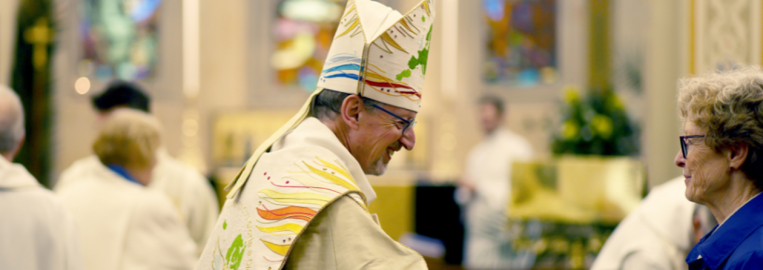 Bishop Robert reaching out to shake hands with a lady in a blue coat in church