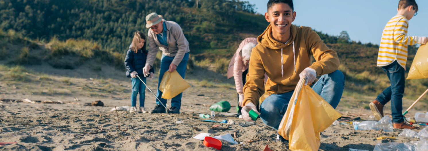 a boy smailes to camera as he puts rubbish in a bag. others clean the beach behind him.