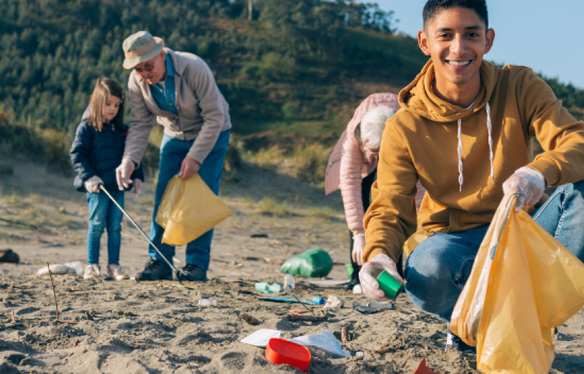 a boy smailes to camera as he puts rubbish in a bag. others clean the beach behind him.
