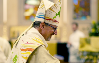 Bishop Robert reaching out to shake hands with a lady in a blue coat in church