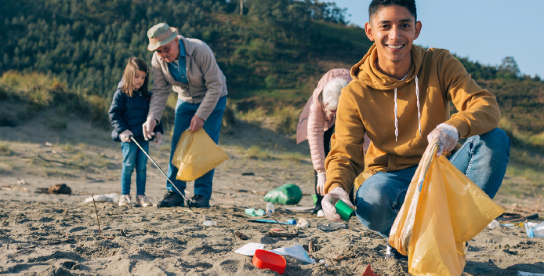 a boy smailes to camera as he puts rubbish in a bag. others clean the beach behind him.